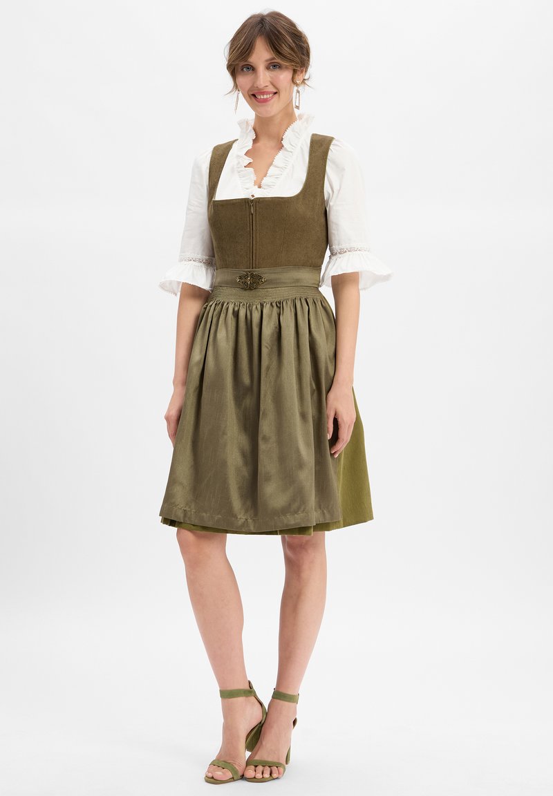 Grünes Kleid mit tailliertem Oberteil, gerafftem Rock und dekorativer Brosche. Weiße Bluse mit Rüschenausschnitt. Olivgrüne Absätze ergänzen das Outfit.