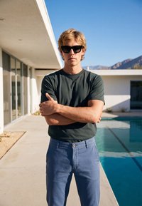 Homme blond portant des lunettes de soleil noires, un t-shirt vert foncé et un pantalon bleu, debout, les bras croisés près d'une piscine devant un bâtiment blanc moderne.