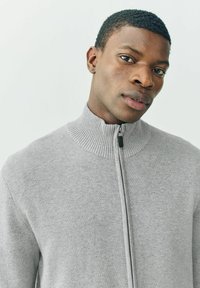 Grauer Strick-Zipfelpullover mit hohem geripptem Kragen und glatter Textur. Verfügt über einen Frontreißverschluss mit einem kontrastierenden schwarzen Zug.