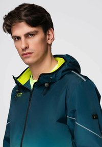 Mladý muž s tmavými vlasy, oblečený v tmavě modré zipové bundě s kapucí a neonově žlutou podšívkou, stojící čelem s neutrálním výrazem.