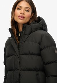 Zwarte donsjas met capuchon, voorzien van een doorgestikt ontwerp, ritssluiting aan de voorkant, drukknopen en een logo patch op de linker mouw. Gladde textuur.