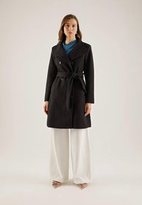 Trench-coat noir à double boutonnage avec ceinture, doté de gros boutons et d'un large col. Associé à un pantalon blanc à jambes larges.