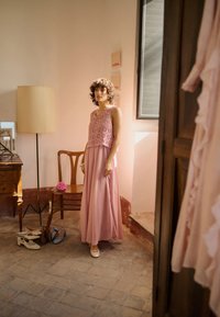 Femme en robe rose sans manches se tient près d'une chaise en bois et d'une table avec plusieurs chaussures sur un sol carrelé dans une pièce doucement éclairée avec une fenêtre.