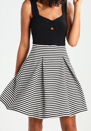 Frau trägt ein ärmelloses schwarzes Oberteil mit Ausschnitt und einen knielangen ausgestellten Rock mit schwarz-weißen horizontalen Streifen.