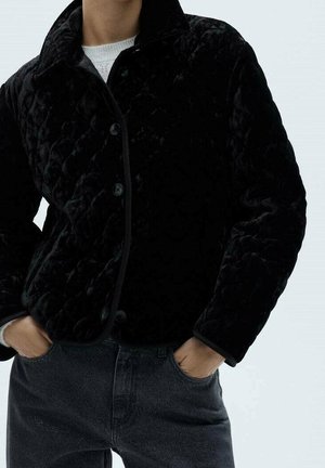 Persoon draagt een zwart gewatteerd jack met knopen, handen in de zakken en donkere spijkerbroek, zichtbaar van nek tot heupen tegen een effen achtergrond.