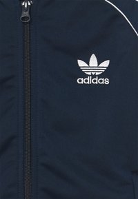 Tmavě modrá bunda Adidas s předním zipem, s bílým vyšívaným logem a kontrastním lemováním podél ramen. Hladká textura.