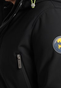 Veste noire à capuche en tissu lisse, avec un cordon jaune en accent, une poche latérale zippée et un écusson rond avec logo sur la manche.