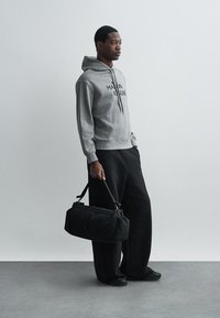 Hanorac gri cu glugă și text negru, pantaloni evazați negri și geantă tip duffel neagră. Material neted și croială lejeră pe toată suprafața.