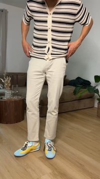 Personne portant une chemise rayée à manches courtes boutonnée, un pantalon beige clair, et des baskets bleu-jaune, debout à l'intérieur sur un sol en bois.