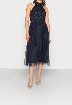 Vrouw draagt een mouwloze marineblauwe halterjurk met een met kraaltjes bezette lijfje en geplooide tule midi-rok, gecombineerd met zilveren openhiel sandalen.