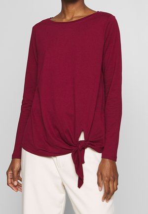 Osoba nosząca bordową bluzkę z długim rękawem, z wiązaniem z przodu, w połączeniu z białymi spodniami na jednolitym tle.