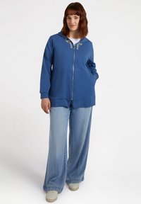 Niebieska bluza z kapturem zapinana na zamek błyskawiczny z dekoracyjnymi akcentami wokół kołnierza, w połączeniu z jasnoniebieskimi spodniami typu wide-leg i jasnymi trampkami.