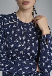 Top a maniche lunghe blu navy con motivo floreale bianco e pois. Presenta un collo rotondo, una chiusura con bottoni e polsini con bottoni e aperture.