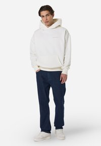 Λευκή φούτερ με μπροστινή τσέπη και ριμπ μανσέτες, συνδυασμένη με σκούρα μπλε τζιν παντελόνι και λευκά αθλητικά παπούτσια. Απλός και άνετος σχεδιασμός.