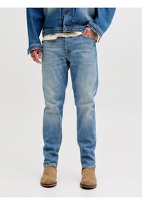 Άτομο που φοράει ανοιχτό μπλε ξεβαμμένα τζιν, καφέ σουέντ μπότες, ένα τζιν μπουφάν και μια μπεζ μπλούζα με το ένα χέρι στην τσέπη.