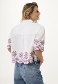 Blusa bianca corta con maniche corte, caratterizzata da un intricato ricamo viola e un design con orlo a scallop. Sul retro c'è un piccolo etichetta.