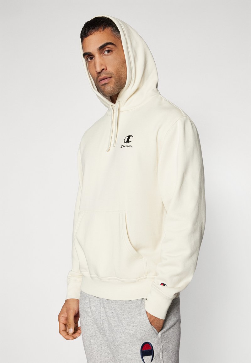 Sudadera con capucha color crema con un bolsillo canguro frontal, logo negro en el pecho y puños acanalados. Llevada con pantalones grises que tienen un parche de logo.