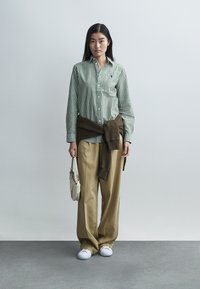 Grün-weiß gestreiftes Hemd, brauner Pullover um die Taille gebunden, beige Hosen mit weitem Bein, weiße Turnschuhe, hält eine kleine Tasche. Einfaches, lässiges Outfit.