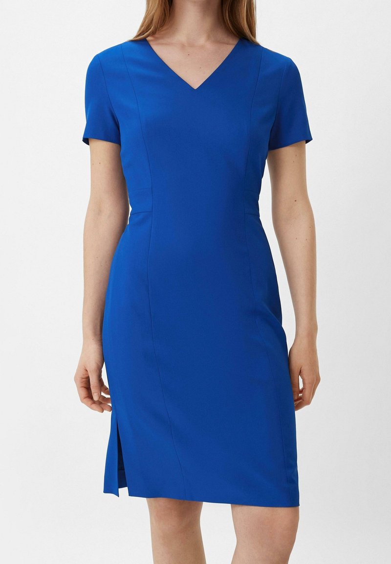 Femme portant une robe bleue ajustée à manches courtes avec un col en V et une fente latérale, debout devant un fond blanc uni.