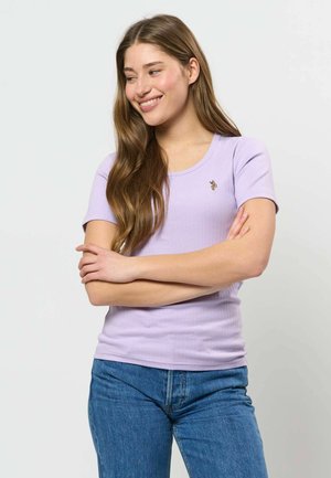 Ung kvinde med langt brunt hår smiler, iført en lyse lilla kortærmet top og blå jeans, armene krydset foran en hvid baggrund.