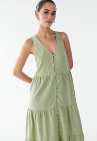 Lichtgroene mouwloze jurk met een V-hals, knoopdetail aan de voorkant en een gelaagd ontwerp van een zachte, getextureerde stof.