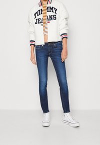 Chaqueta bomber blanca con detalles en azul marino y rojo y logo de "TOMMY JEANS", top de rayas recortado, jeans ajustados azules y zapatillas altas blancas.