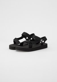 Svarta sandaler med vävda tygremmar, en platt gummisula och en vadderad fotbädd. Designen inkluderar korsremmar och en märkesdetalj.