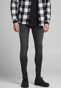 Hombre vestido con una camisa a cuadros en blanco y negro, camiseta negra, jeans oscuros ajustados, zapatos de vestir negros y anillos en ambas manos.