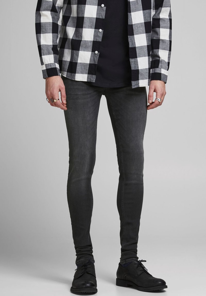 Hombre vestido con una camisa a cuadros en blanco y negro, camiseta negra, jeans oscuros ajustados, zapatos de vestir negros y anillos en ambas manos.