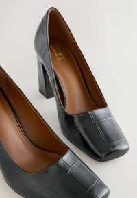 Czarne lakierowane szpilki ze kwadratowym noskiem, fakturą imitującą krokodyla oraz masywnym, szerokim obcasem, z brązową podszewką wewnętrzną.