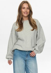 Svetlo siva oversized dukserica s spuštenim ramenima i rebrastim manžetama, uparena sa visokim plavim farmerkama. Glatka tekstura, ležeran dizajn.