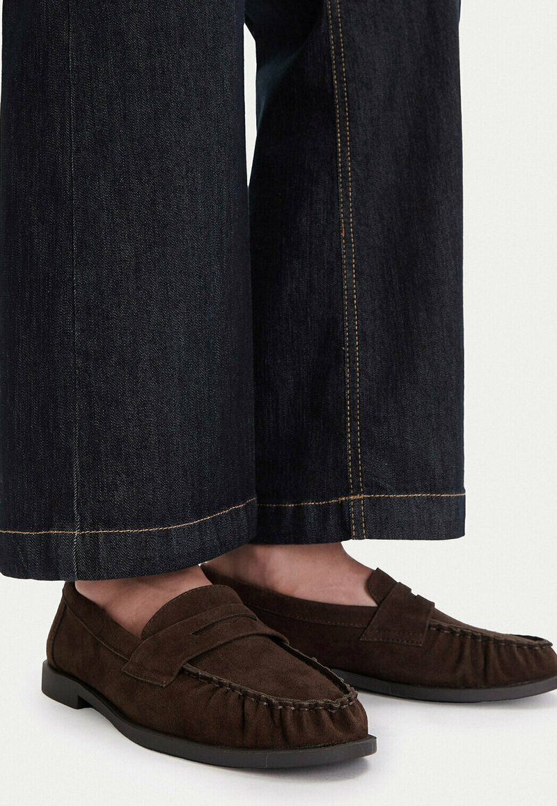 Mocasines de ante marrón oscuro usados con vaqueros de pierna ancha en tono oscuro, mostrando costuras detalladas en los zapatos y los dobladillos.