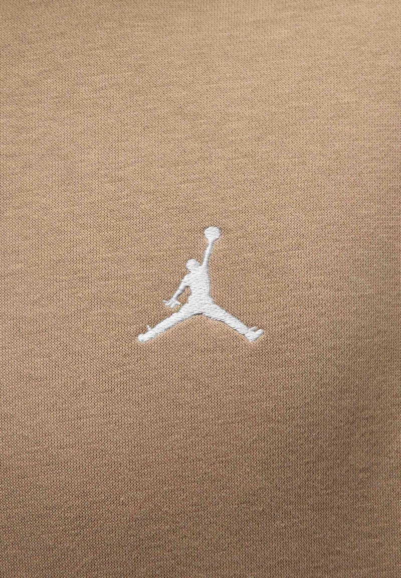 Logo di marca su tessuto color beige; presenta un intricato ricamo bianco di una figura che salta tenendo una pallacanestro. Texture morbida, tessuto misto.