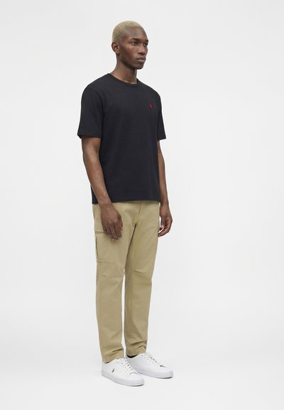 Νέος άνδρας με κοντά ξανθά μαλλιά, φορώντας μαύρο μπλουζάκι, μπεζ παντελόνι cargo και λευκά αθλητικά, στέκεται μπροστά σε μια απλή λευκή φόντο.