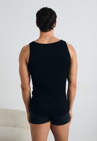 Schwarzes ärmelloses Tanktop aus geripptem Stoff, mit rundem Ausschnitt und enganliegendem Design. Getragen mit dunklen Shorts.