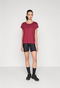 T-shirt corto color marrone bordeaux con una vestibilità morbida, abbinato a pantaloni ciclistici neri con texture. Il modello indossa scarpe da ginnastica nere e calze fino al ginocchio.