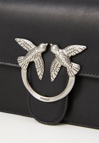 Sac en cuir noir avec un accent en métal argenté circulaire ornée de deux sculptures d'oiseaux, détaillées avec des plumes texturées. Marque : Pinko.