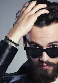 Ochelari de soare negri cu accente portocalii, o geacă din piele neagră și o brățară din mărgele închise. Subiectul are părul întunecat de lungime medie și o barbă.
