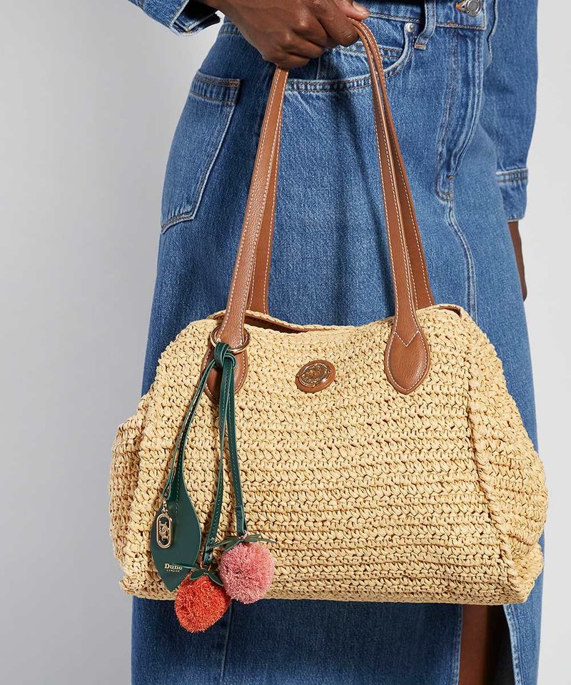 Main tenant un sac à main tressé beige avec des anses en cuir marron et des breloques pompons colorées, sur un fond de robe en jean bleu.