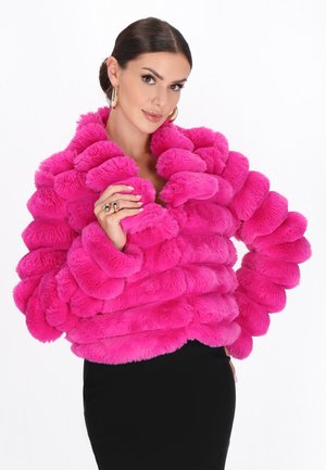 Vrouw met donker haar in een strakke knot, gekleed in grote gouden hoepeloorbellen en een felroze pluizige bontjas, poseert met een hand dicht bij haar gezicht.