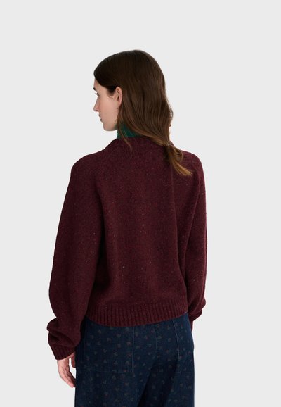 Femme aux cheveux bruns en queue de cheval basse, portant un pull rouge foncé et un pantalon bleu avec un motif subtil, vue de dos sur un fond uni.