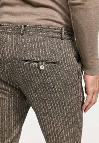 Braun gemusterte Hose mit subtilen Streifen, mit einer geknöpften Gesäßtasche und Gürtelschlaufen, teilweise sichtbar gestecktes Hemd.