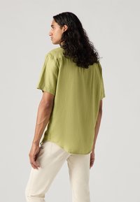 Camicia a maniche corte in tessuto verde oliva chiaro, vestibilità rilassata, leggermente strutturata con orlo curvo; abbinata a pantaloni bianco sporco.