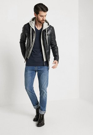 Veste en cuir noir avec une capuche grise, t-shirt bleu marine à col en V, jean bleu clair et bottines noires à la cheville. Modèle dans une pose debout décontractée.