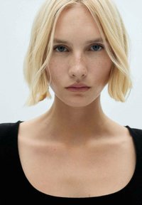 Portretul unei persoane cu păr scurt, blond și ochi albaștri, purtând un top negru. Caracteristicile includ pistrui deschisi și o expresie neutră.
