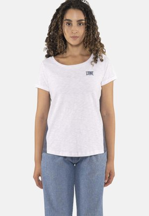 Mujer con cabello rizado que lleva una camiseta blanca de manga corta con el logo "LEONE" y vaqueros azules, mirando hacia adelante sobre un fondo claro.