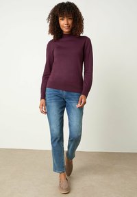 Lila Langarmpullover mit hohem Kragen, kombiniert mit blauen Jeans. Die Schuhe sind braune Wildleder-slipper. Neutrale Hintergrund.