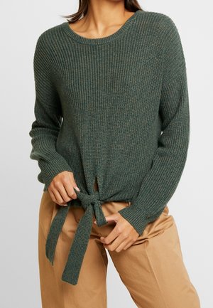Femme portant un pull en maille côtelée vert foncé avec un lien à nouer à la taille et un pantalon beige, mains ajustant le lien.
