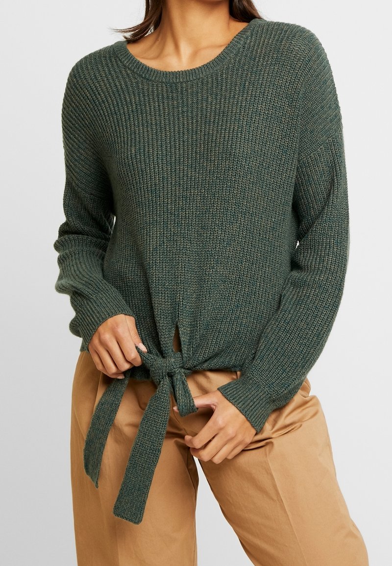 Femme portant un pull en maille côtelée vert foncé avec un lien à nouer à la taille et un pantalon beige, mains ajustant le lien.