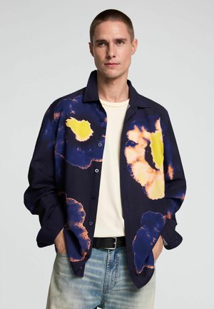 Mann trägt dunkelblaues Hemd mit Knöpfen mit großen gelben und orangefarbenen abstrakten Mustern, cremefarbenes T-Shirt und hellblaue Jeans, Hände in den Taschen.
